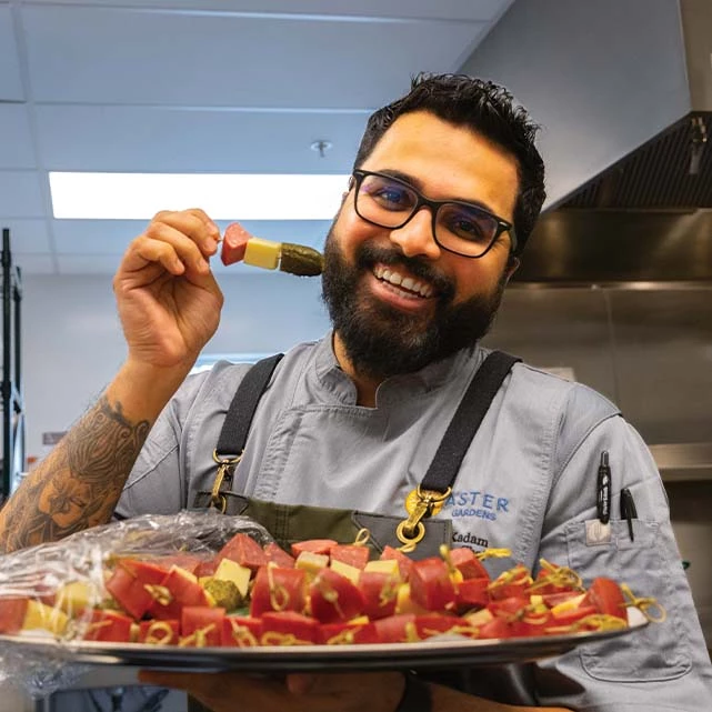 Aster Gardens chef smiling while holding a plate of appetizers including pickles, cheese, and meat.