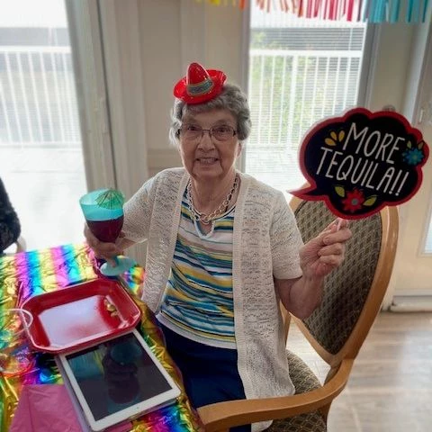 A senior resident posing with Cinco de Mayo decorations. She is holding a drink and a sign that says,