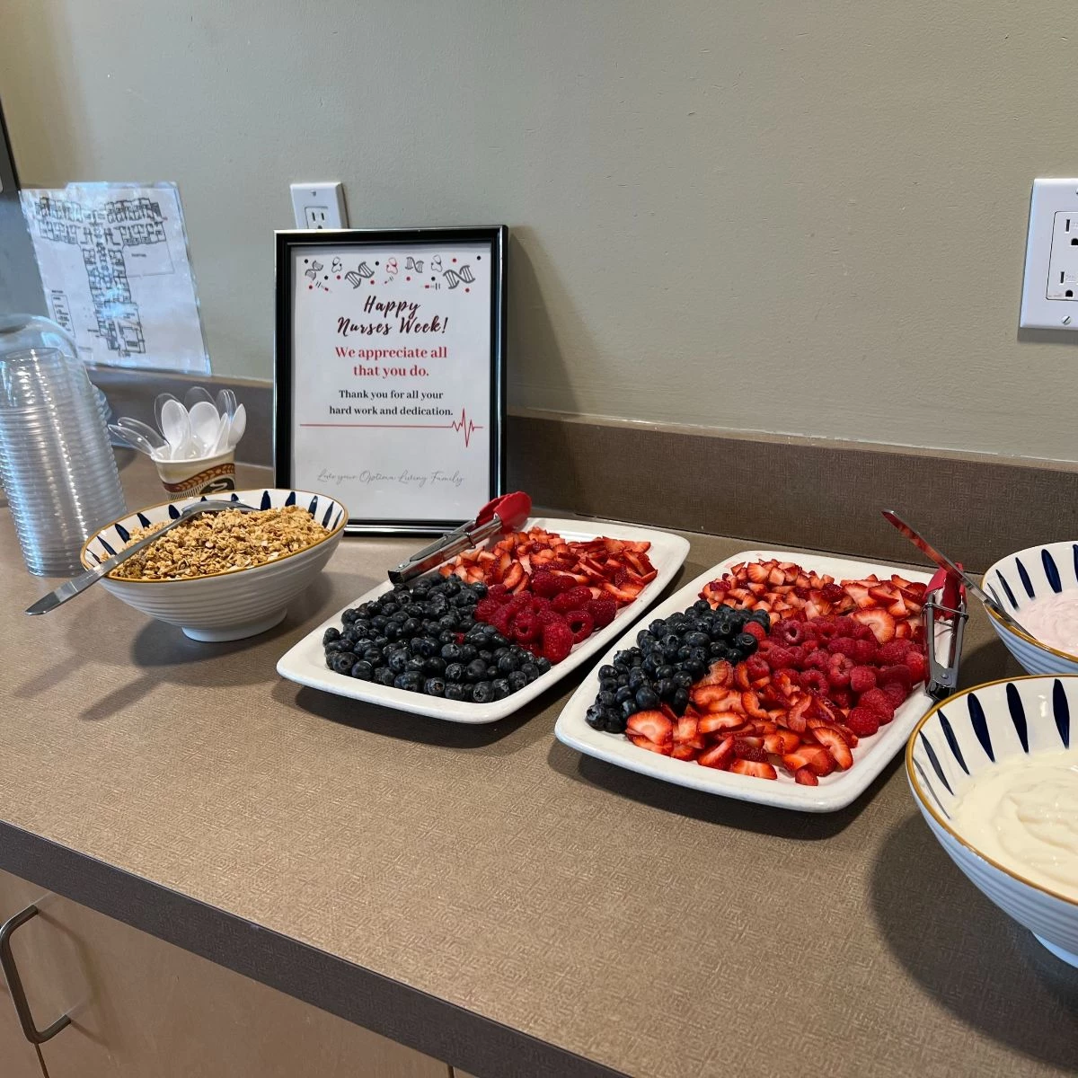 Some platters of fruit along with yogurt and granola available to be served. Behind is a sign that says,