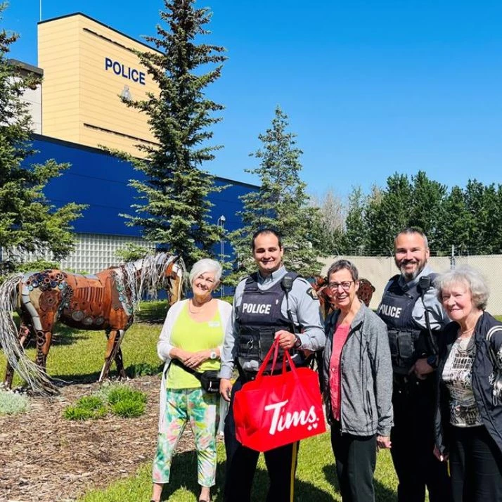 Seniors standing with some police officers in front of a police station. The one officer is holding a bag of Tim Hortons food.