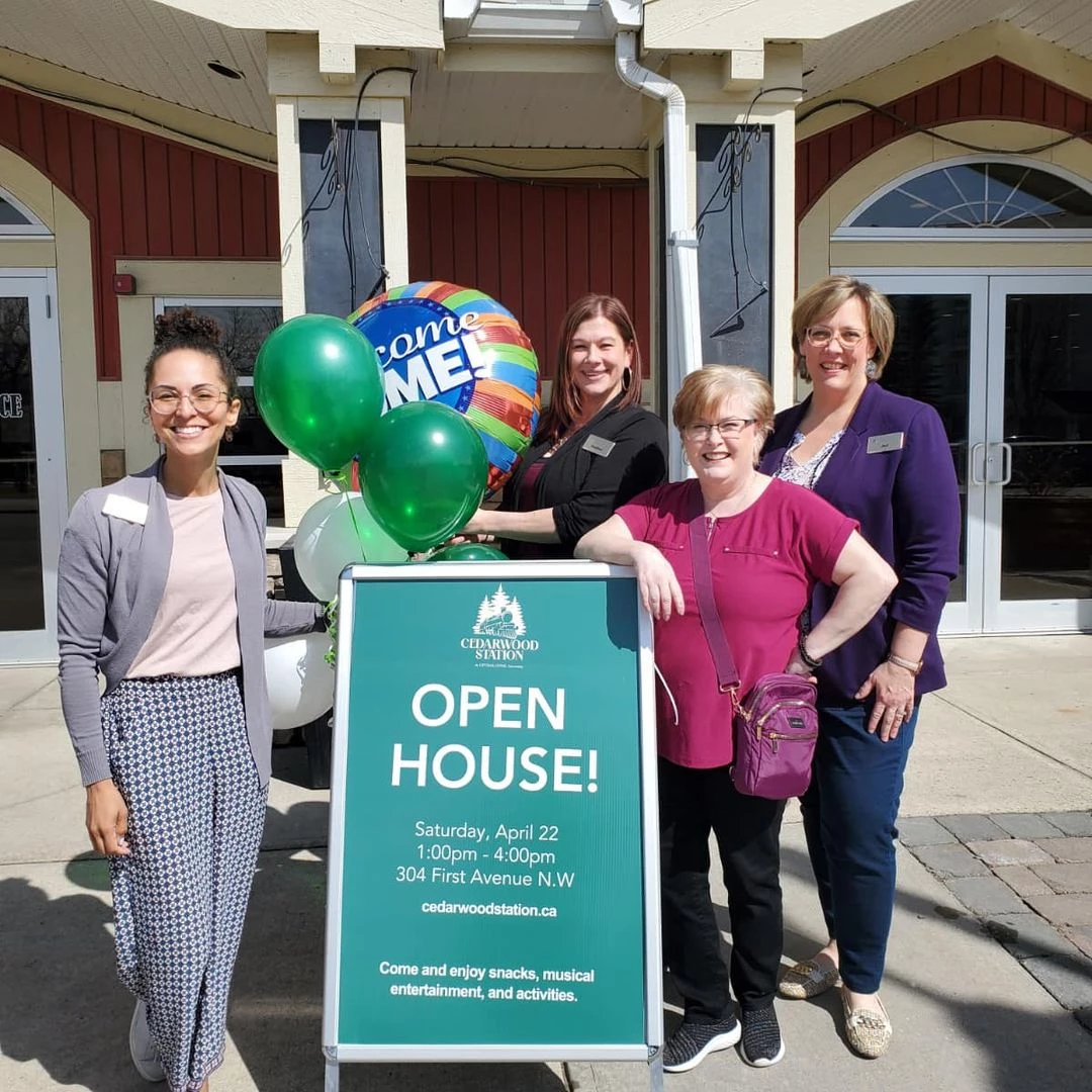 Four staff members standing together with balloons and an open house sign. The balloons are mostly green and white, along with a large balloon that says,